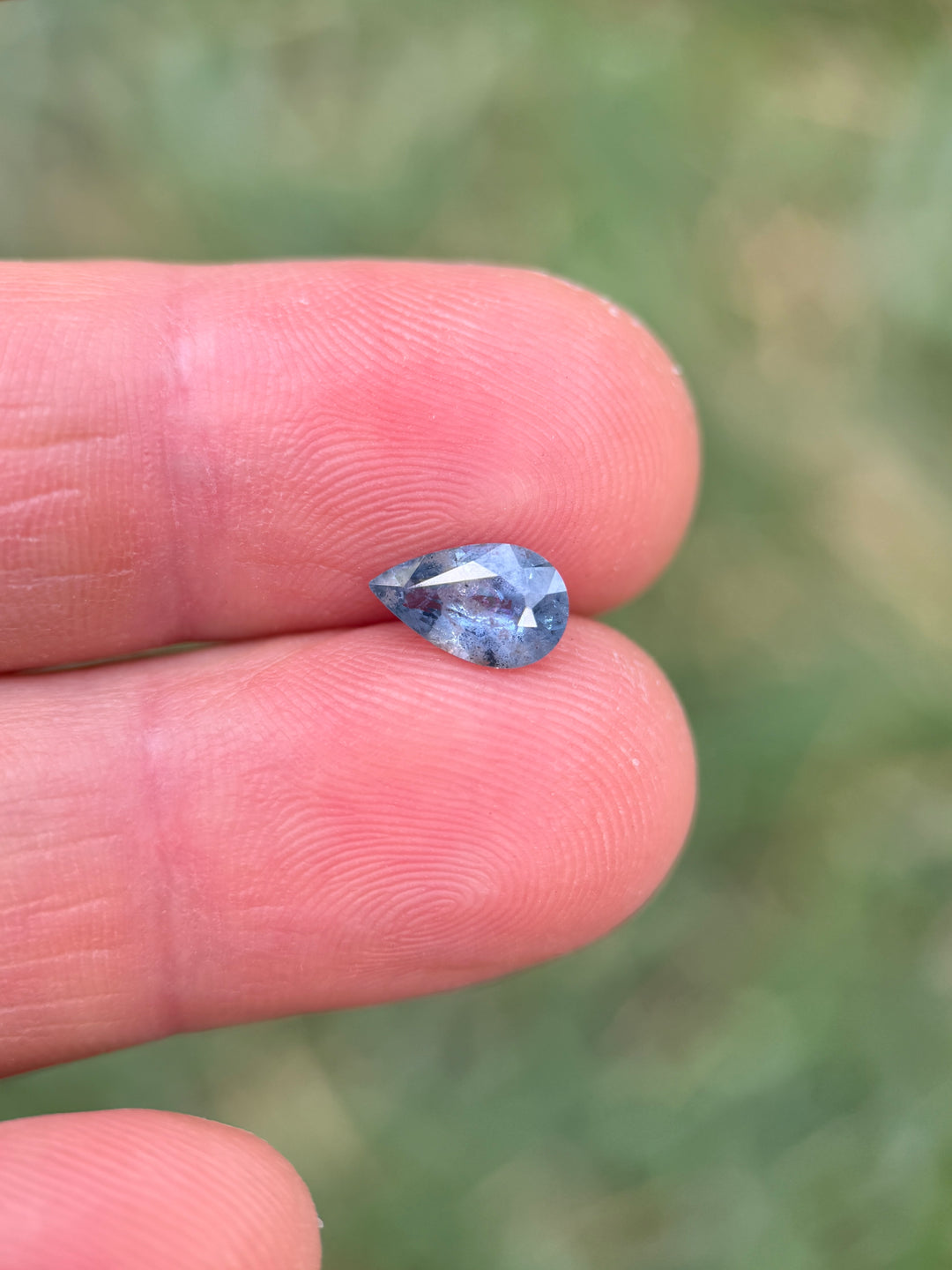 1.30 carat periwinkle blue sapphire balanced on fingertips for scale, revealing its pear mix cut shape and soft blue glow, natural and untreated gem from the Umba River Valley, Tanga, Tanzania.