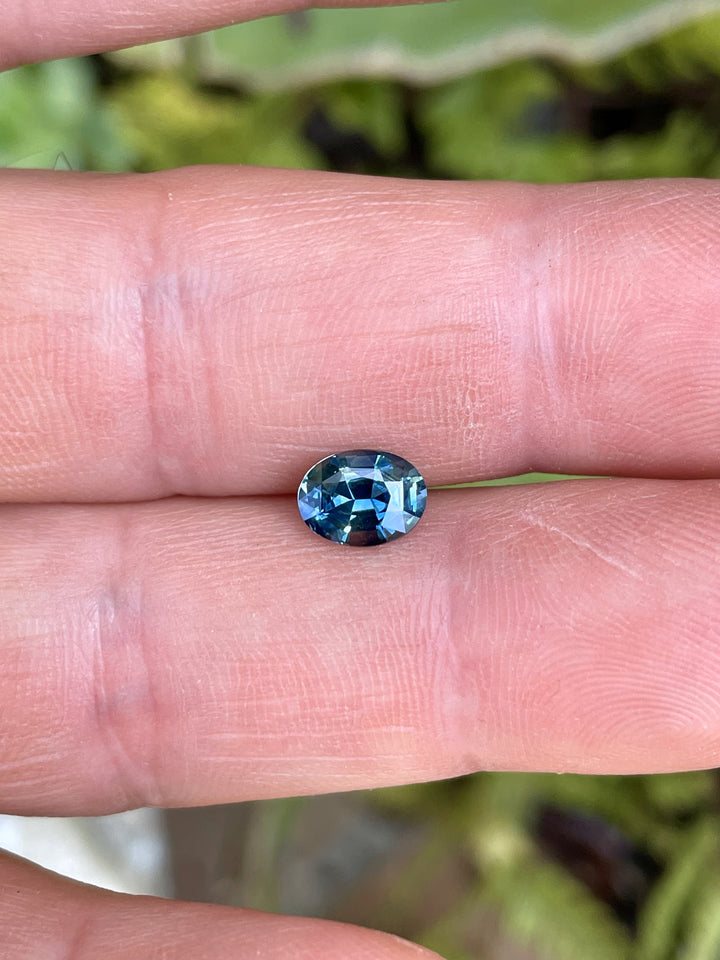 A 1.30-carat mix modified oval brilliant sapphire with teal, blue, and green hues, resting on fingertips under natural light.