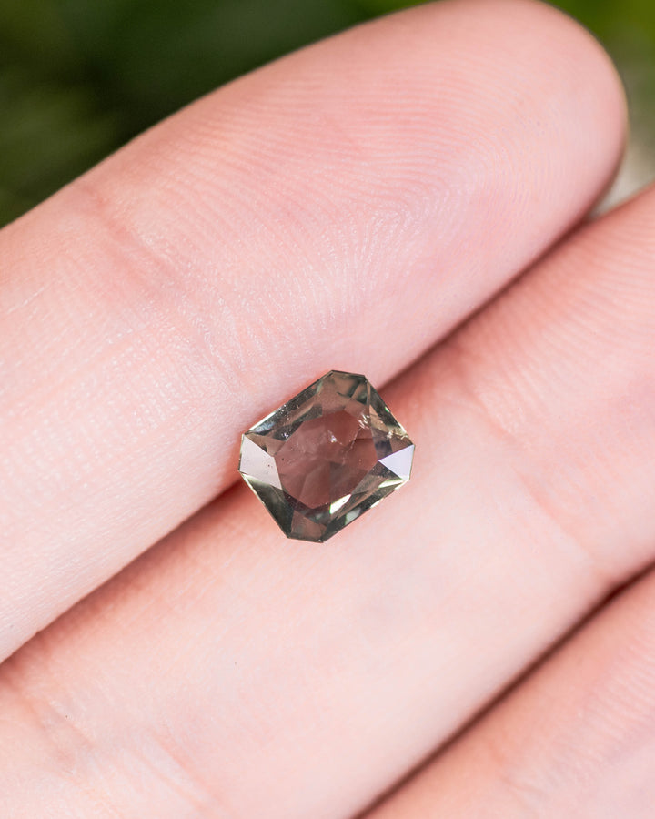 Close-up of 1.31 carat teal, yellow, and green sapphire resting on a fingertip, showing color zoning and brilliance through its custom modified radiant cut, untreated gemstone from Umba River Valley, Tanga, Tanzania.