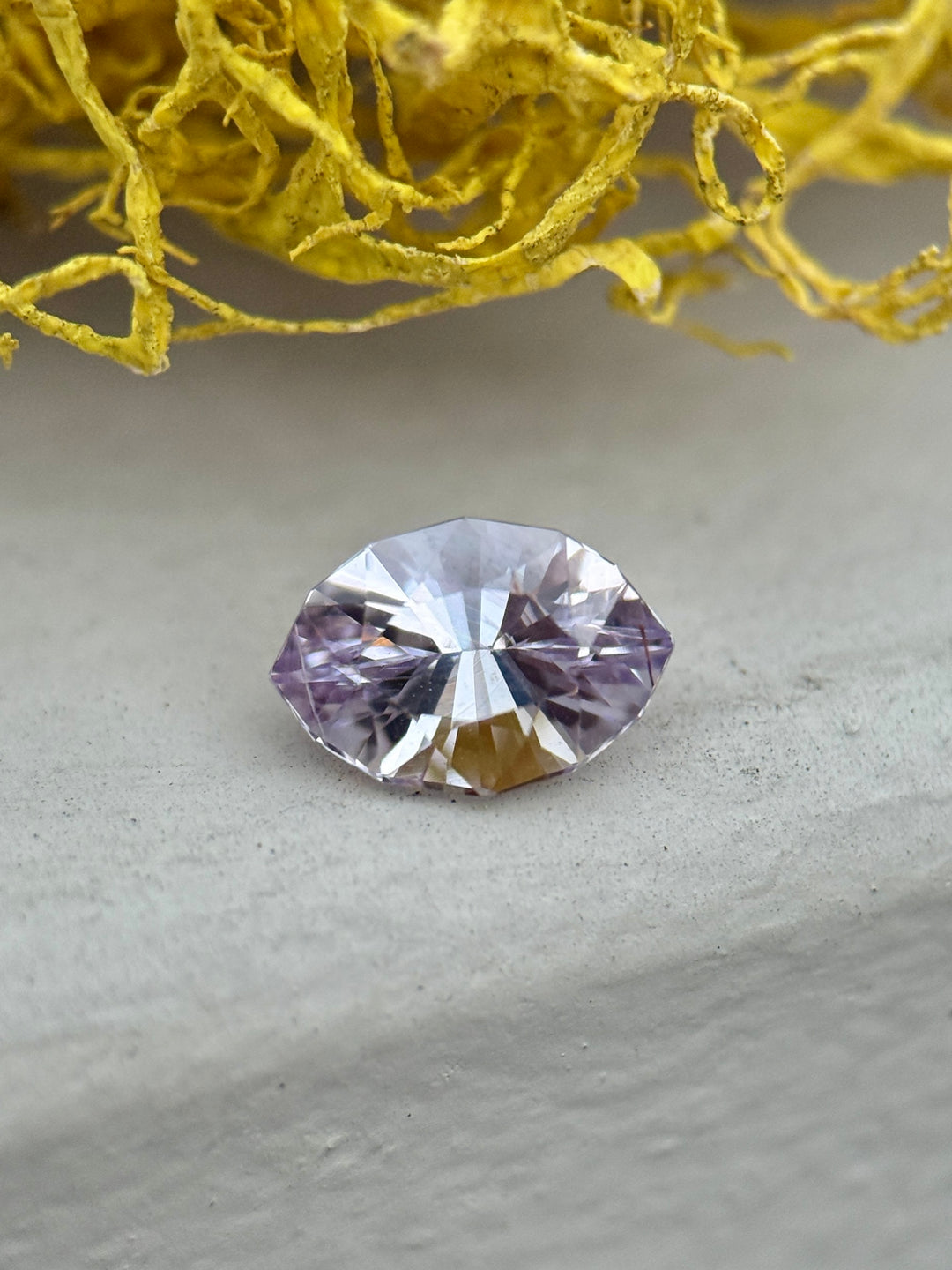 1.32 carat pink, purple, and lilac sapphire resting on a stone surface beside yellow moss, showcasing pastel hues and sparkle in a precision marquise mixed cut, untreated sapphire from Sakaraha, Madagascar.