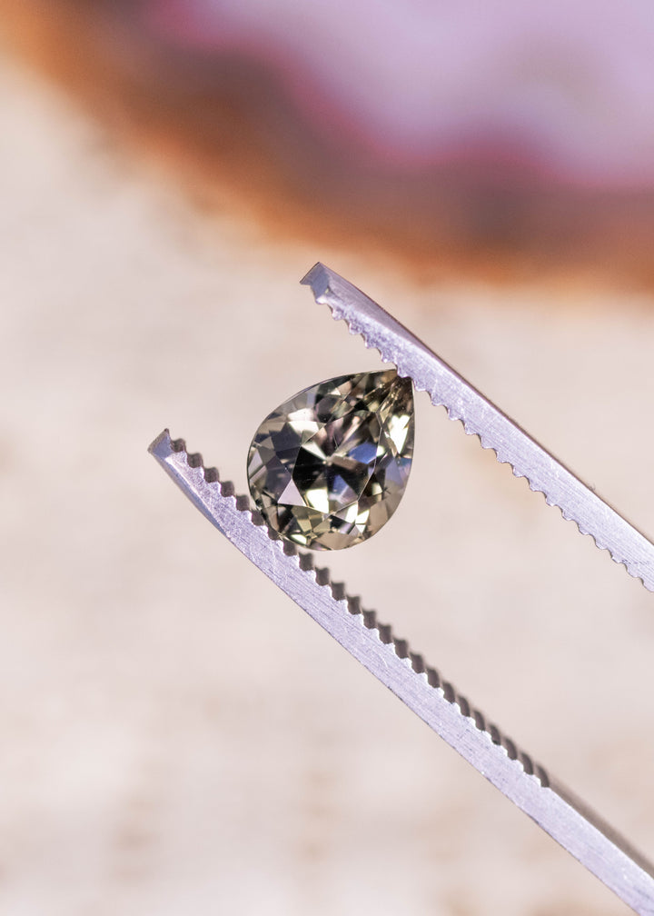 Loose 1.32ct pear cut Tanzanite held in gem tweezers displaying green brown purple tones and brilliant sparkle for custom jewelry design