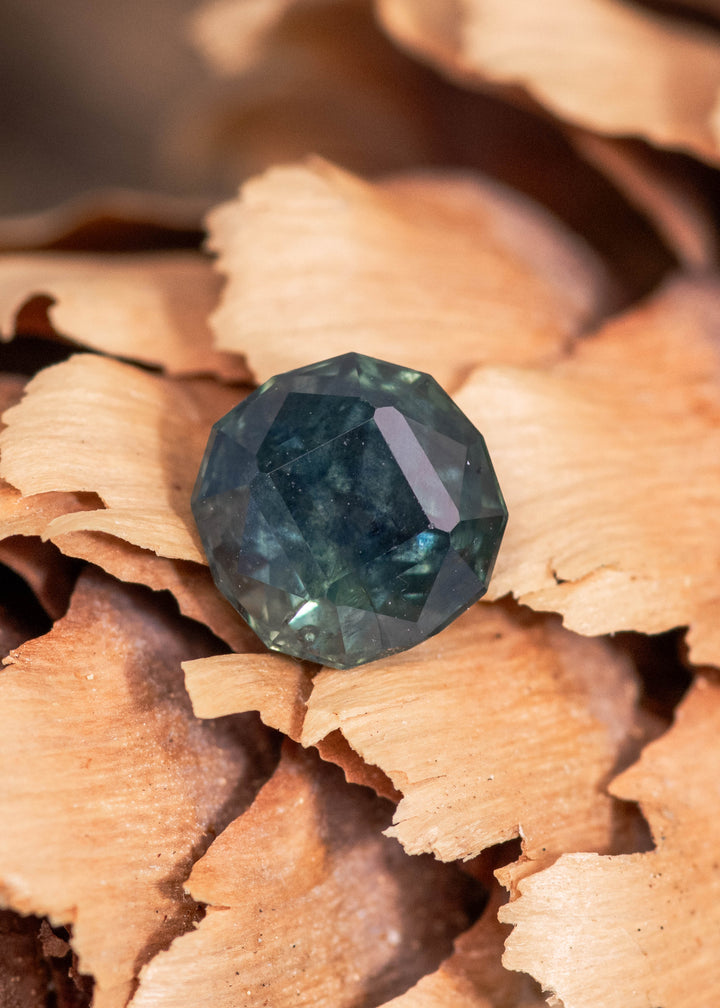 1.32 carat blue green teal sapphire displayed on natural wood texture, unheated Montana gemstone with geometric rounded cushion faceting and deep teal tones