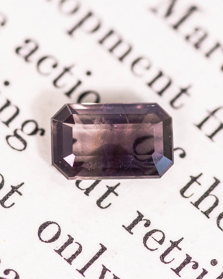 1.34 carat purple, violet, and brown sapphire resting on a vintage book page, showcasing geometric symmetry and depth in its modified emerald cut, untreated gemstone from Umba River Valley, Tanga, Tanzania.