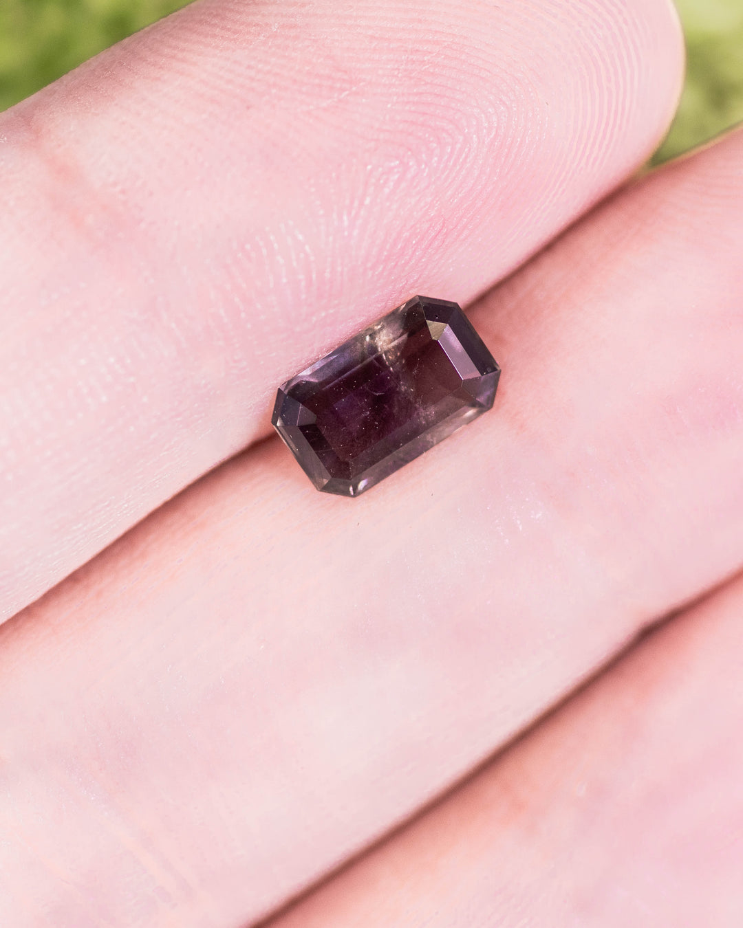Close-up of 1.34 carat purple, violet, and brown sapphire placed on a fingertip, displaying its deep color saturation and clarity in a modified emerald cut, untreated gemstone from Umba River Valley, Tanga, Tanzania.