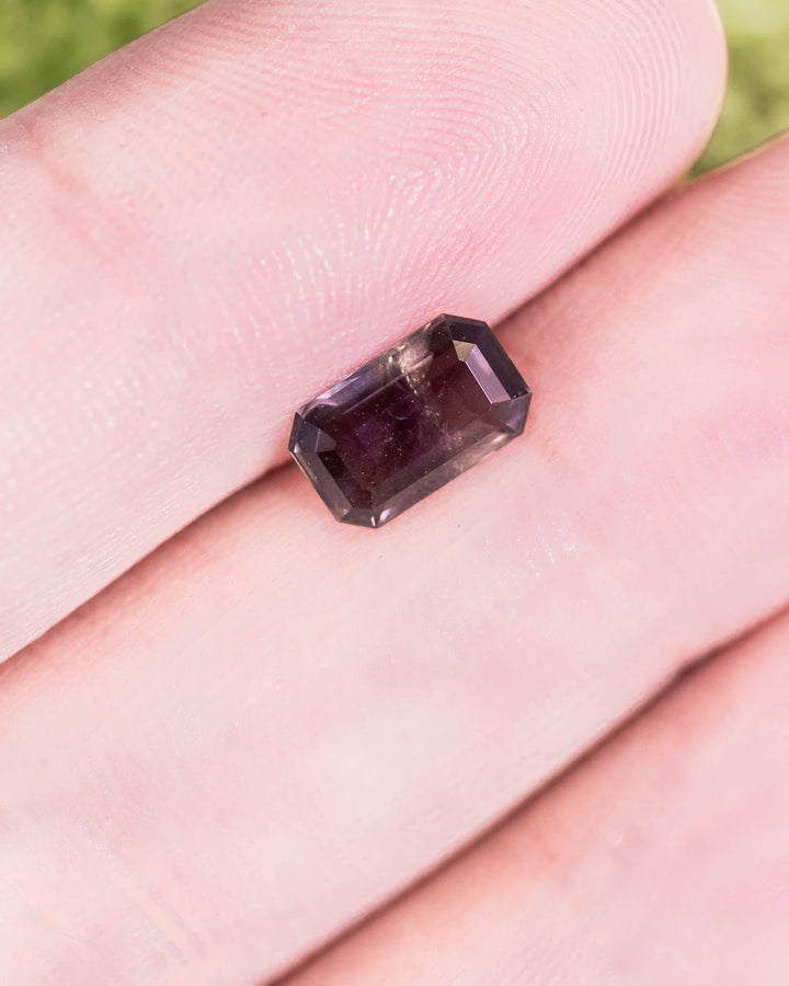 Close-up of 1.34 carat purple, violet, and brown sapphire placed on a fingertip, displaying its deep color saturation and clarity in a modified emerald cut, untreated gemstone from Umba River Valley, Tanga, Tanzania.