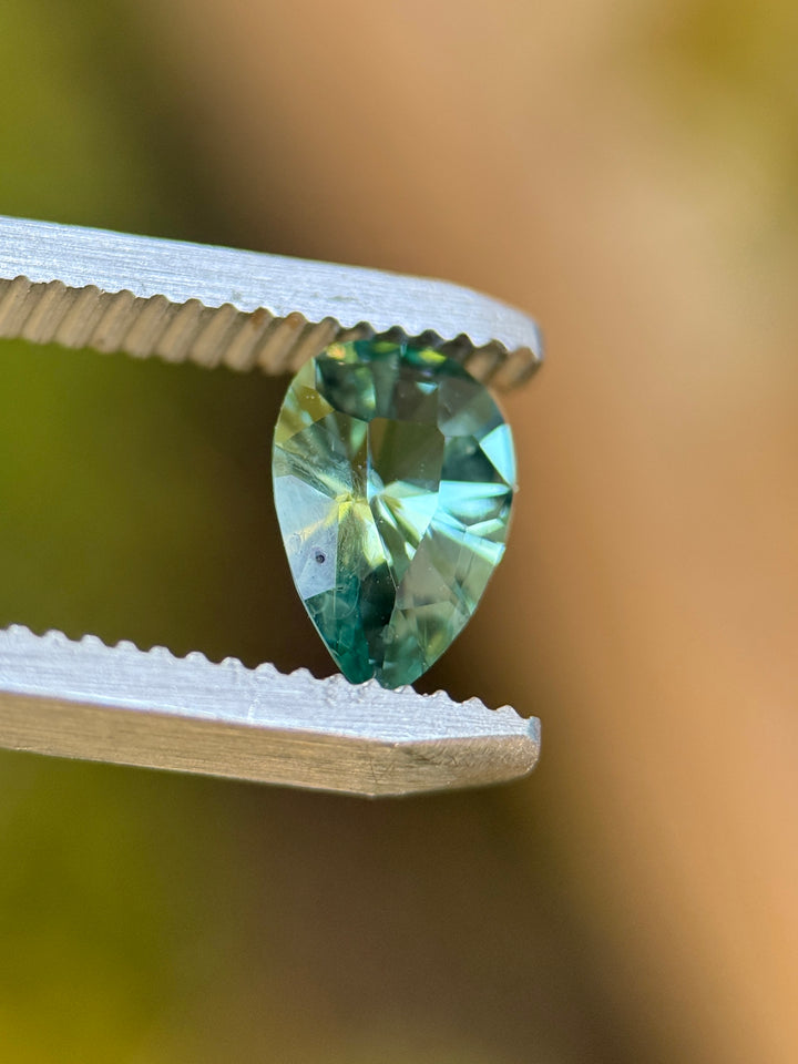Side view of a lagoon teal green Montana sapphire held in gem tweezers, highlighting the shield step cut facets, depth, and heated sapphire clarity.