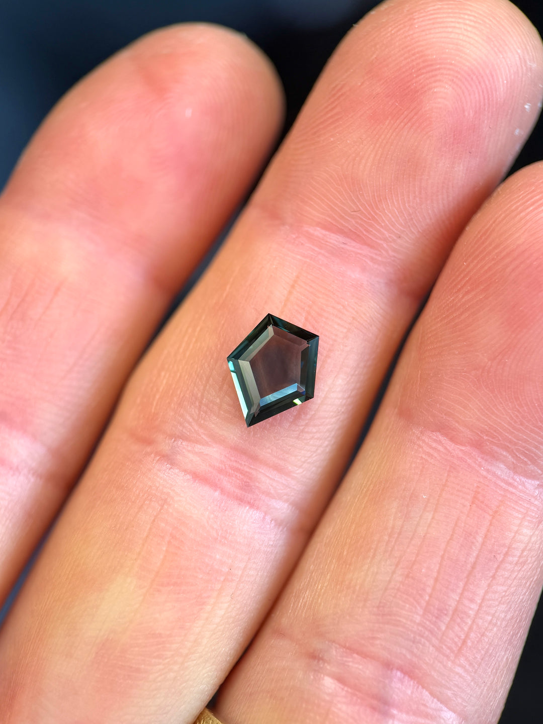 Loose 1.35 carat sapphire resting on fingertips, showing blue, green, and teal tones in a custom shield portrait cut from Gombe, Nigeria.