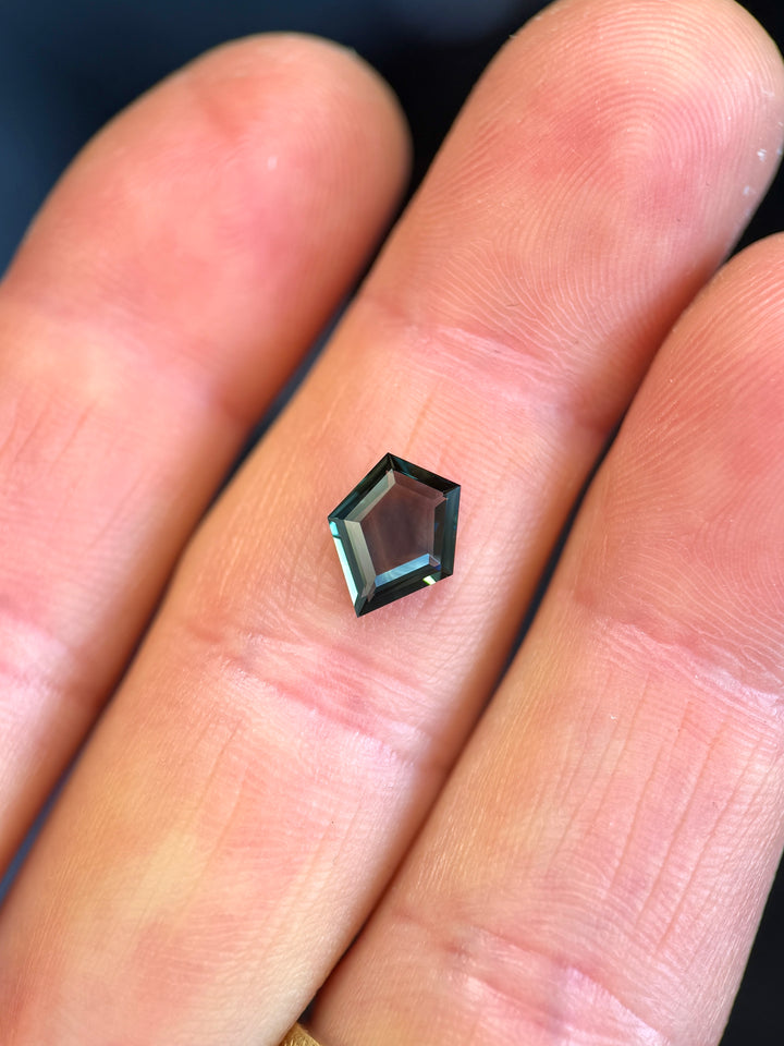 Loose 1.35 carat sapphire resting on fingertips, showing blue, green, and teal tones in a custom shield portrait cut from Gombe, Nigeria.