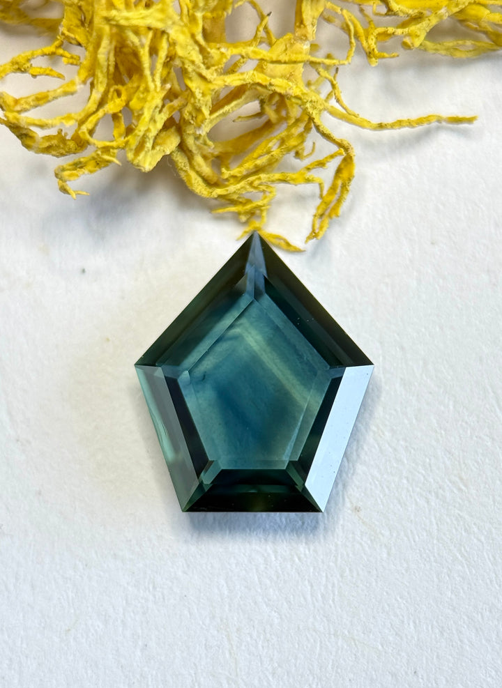 Loose custom shield portrait cut sapphire displayed on a white surface, revealing layered blue, green, and teal color zoning from Nigeria.