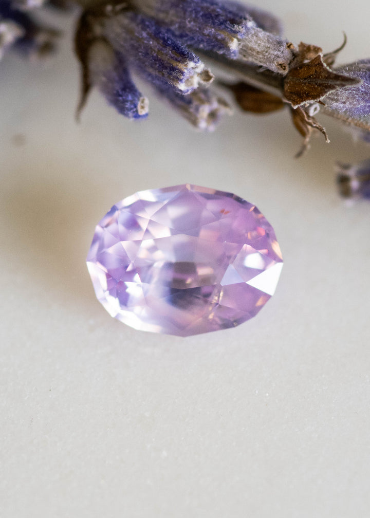 Loose 1.37 carat opalescent pink peach sapphire from Ratnapura shown face-up on a white surface, displaying soft pastel glow and precision oval mix cut facets.