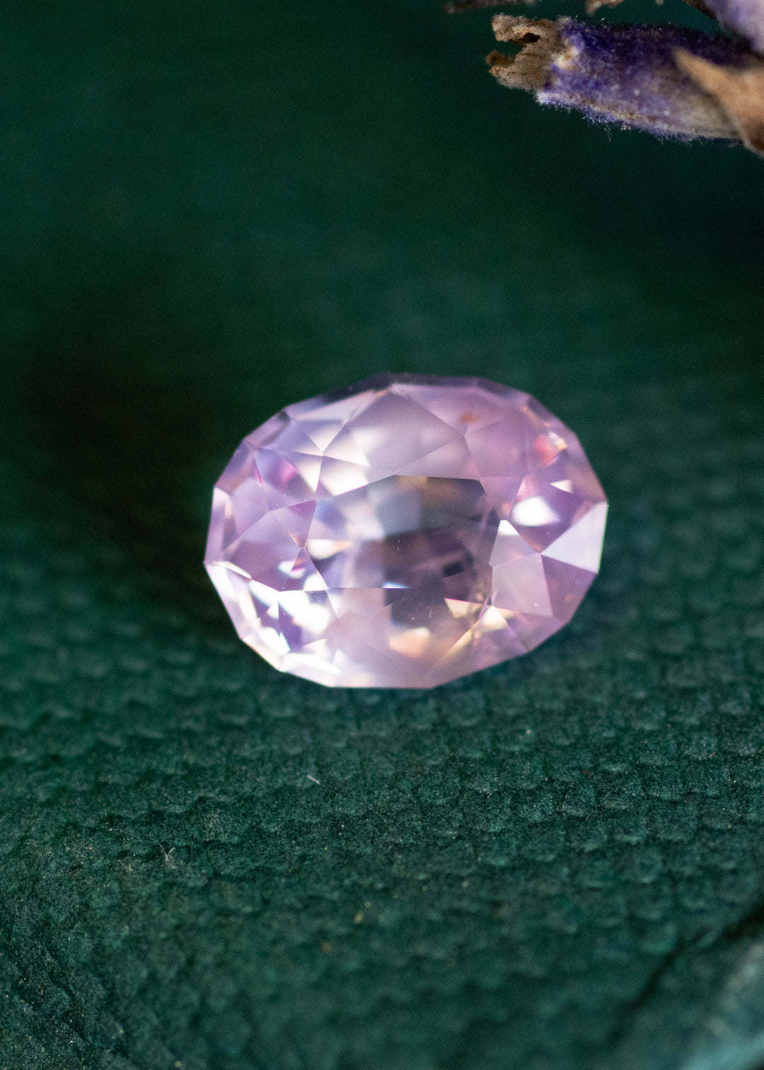 Loose 1.37 carat opalescent pink peach sapphire from Ratnapura shown face-up on a textured green surface, displaying soft pastel glow and precision oval mix cut facets.