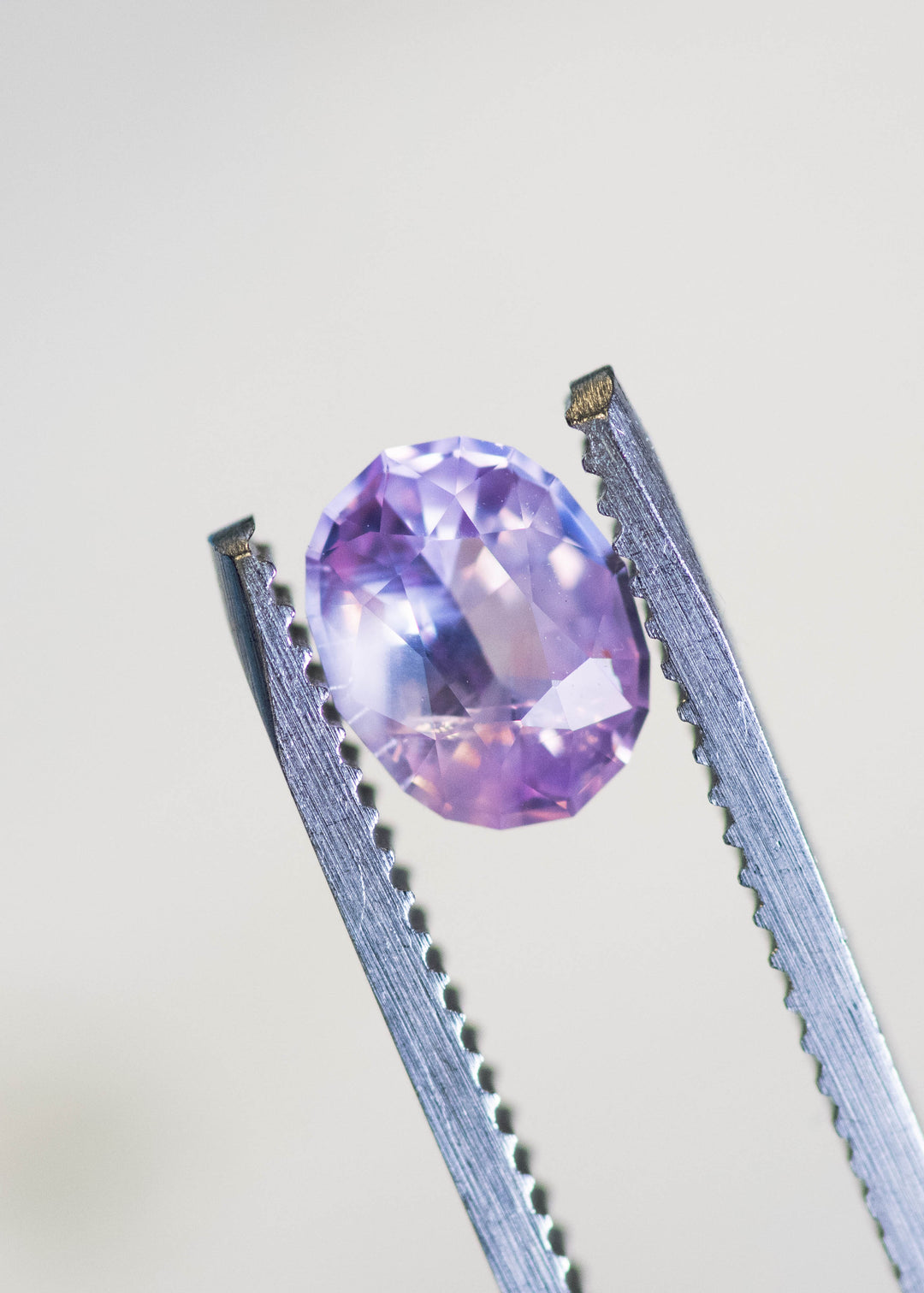 Loose opalescent pink peach sapphire held in tweezers, revealing balanced oval proportions and gentle lavender-peach color shift under natural light.
