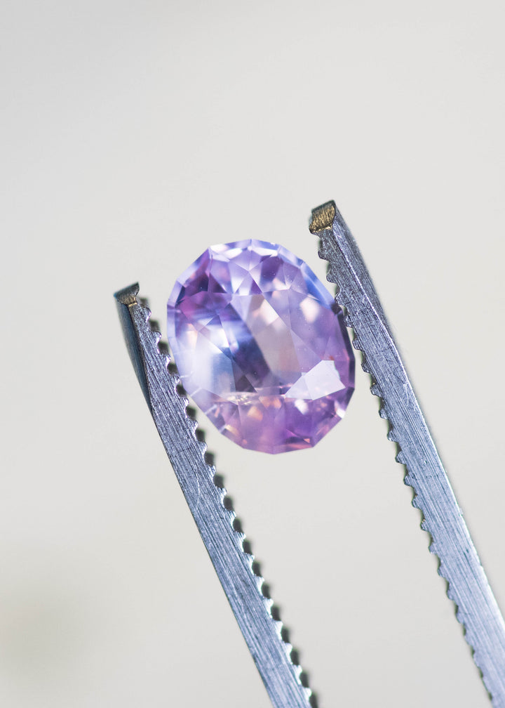 Loose opalescent pink peach sapphire held in tweezers, revealing balanced oval proportions and gentle lavender-peach color shift under natural light.