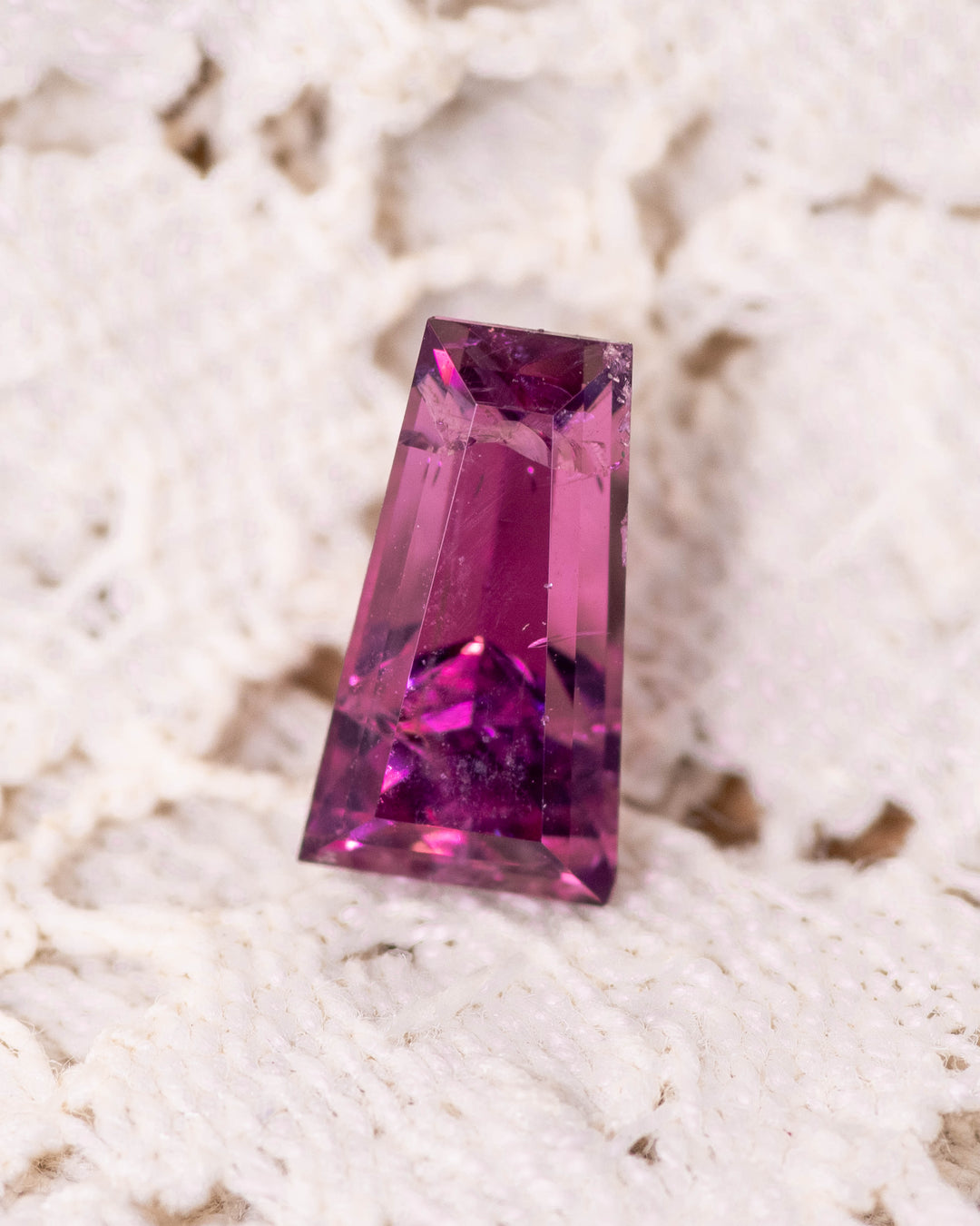 1.39 carat pink and purple garnet resting on a textured ivory fabric, showcasing brilliance, clarity, and sharp symmetry of the step cut tapered baguette, untreated garnet from Kenya.