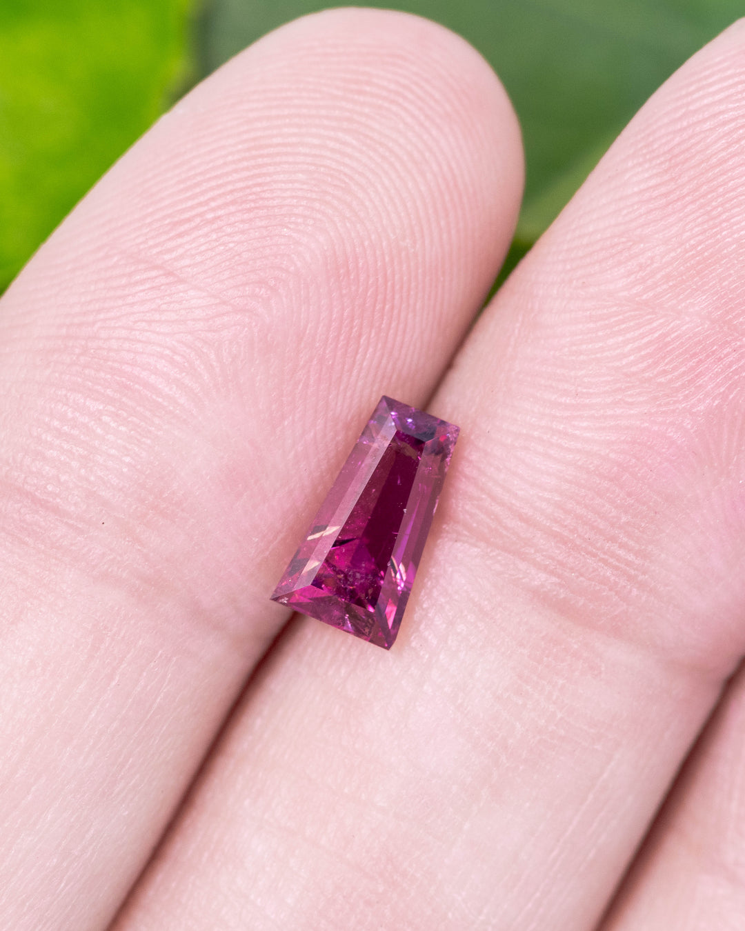1.39 carat pink and purple garnet displayed on fingertips, capturing sparkle, luster, and transparency of the step cut tapered baguette, untreated garnet from Kenya.