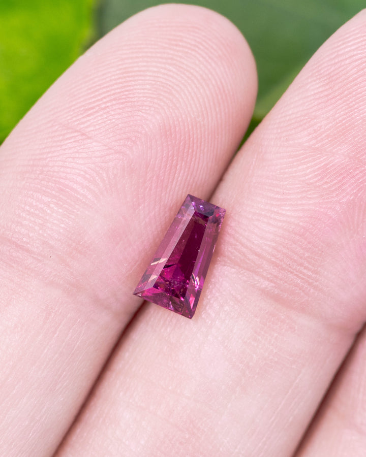 1.39 carat pink and purple garnet displayed on fingertips, capturing sparkle, luster, and transparency of the step cut tapered baguette, untreated garnet from Kenya.