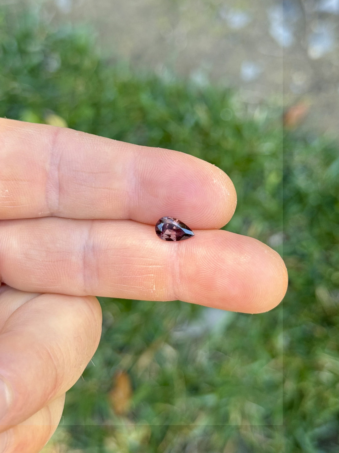 Close-up of a 1.39-carat unheated pear-shaped sapphire with red and purple hues held between fingers, photographed outdoors