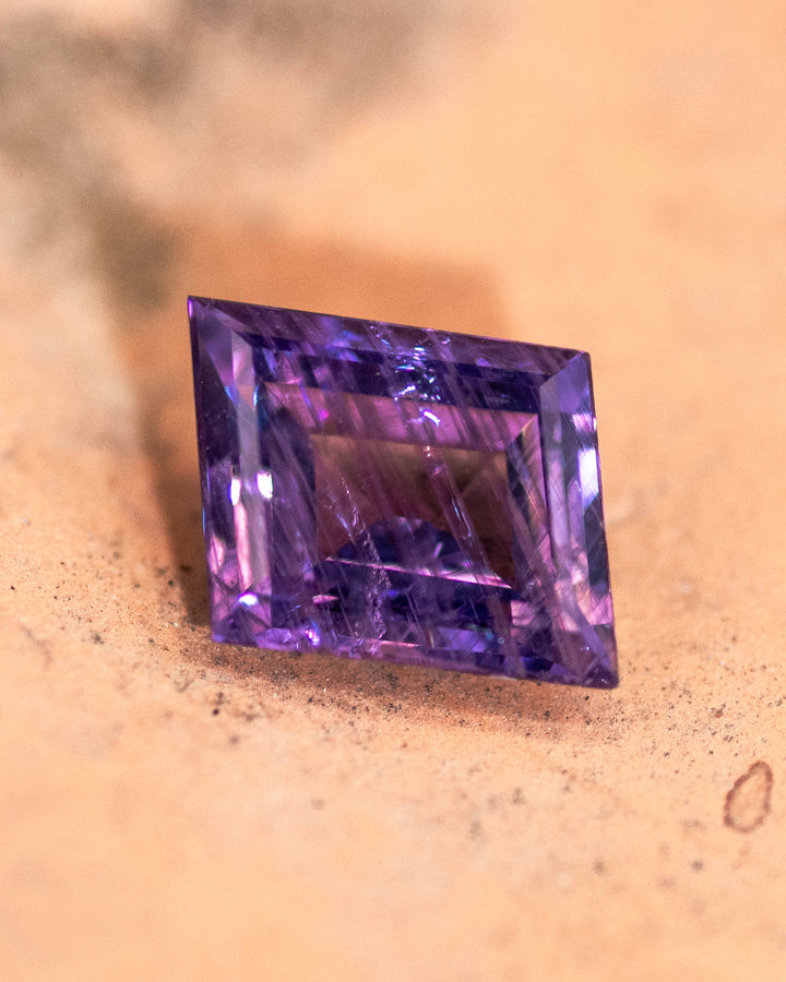 1.40 carat purple sapphire resting on textured tan stone surface, showcasing the vivid hue and geometric facets of the rhombus step cut, untreated gemstone from the Umba River Valley in Tanzania.