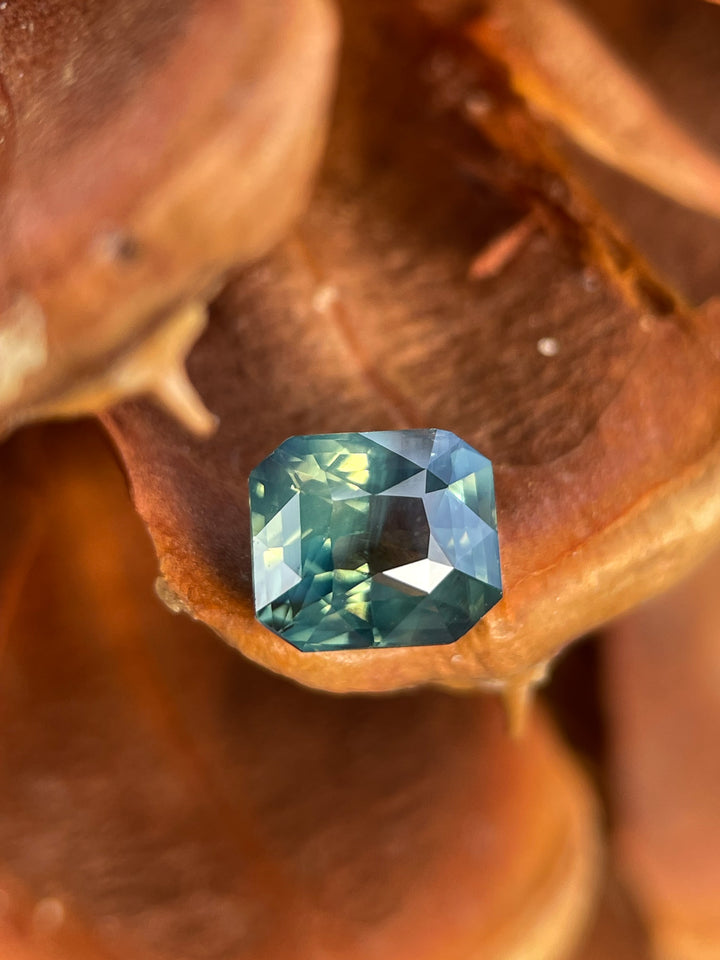 A beautiful 1.42-carat untreated sapphire with opalescent yellow, teal, and green hues. Features a radiant cut, ethically sourced from Madagascar, displayed against a warm natural backdrop.