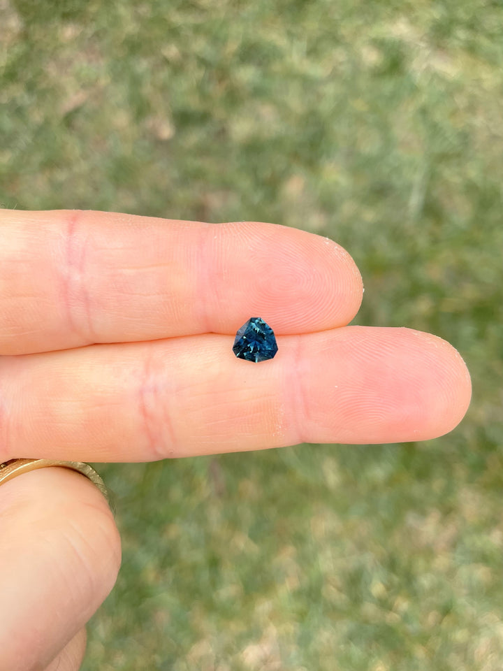A 1.43-carat no-heat sapphire with a striking teal and blue-green hue. The gemstone is precision-cut into a custom geometric trillion shape, displayed on a fingertip to showcase its brilliant facets. Originating from Madagascar, this stone is untreated and priced at $1,072.50.