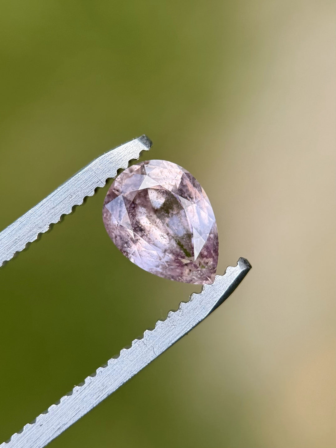Close-up of a 1.45-carat pear-shaped sapphire with blush mauve, peach, and salt and pepper inclusions, held by gem tweezers