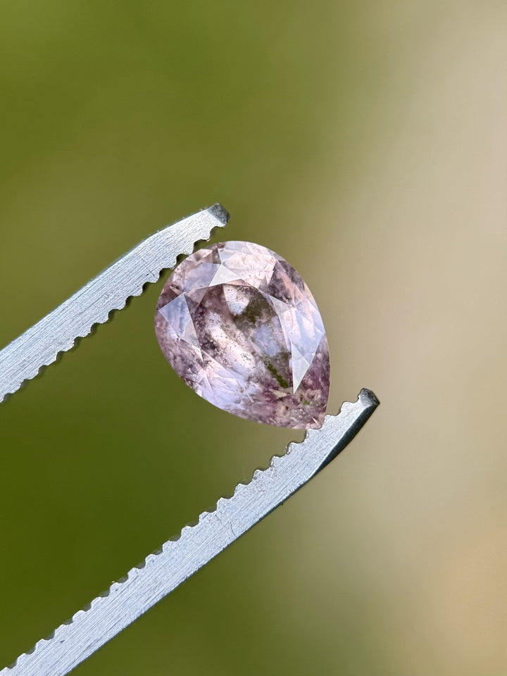 Close-up of a 1.45-carat pear-shaped sapphire with blush mauve, peach, and salt and pepper inclusions, held by gem tweezers