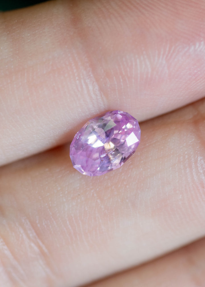 Close-up of 1.50ct lavender pink sapphire oval cut displaying soft opalescent glow resting on finger tips.