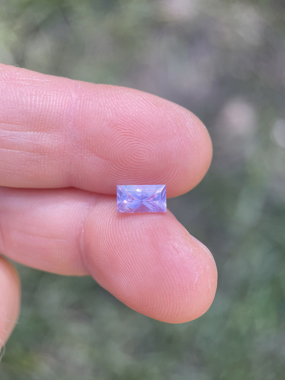 Handheld view of a 1.53-carat baguette-cut sapphire from Sri Lanka, revealing opalescent pink and purple hues in natural light.