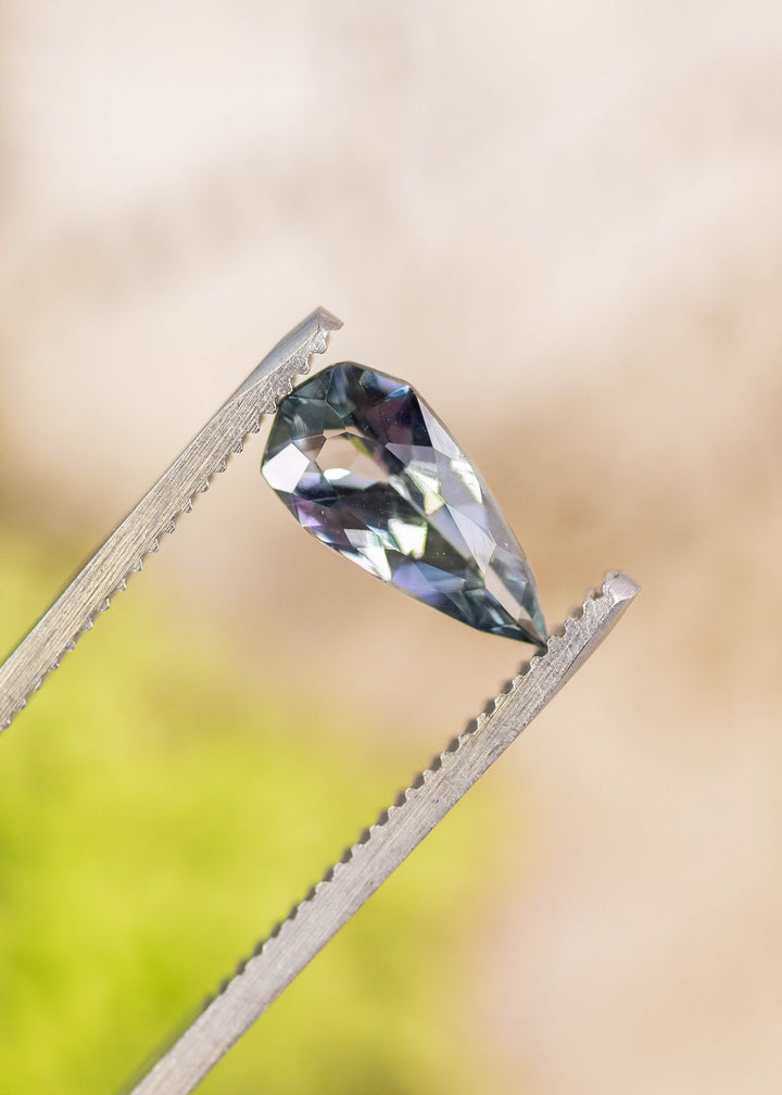 1.53 carat blue teal violet Tanzanite custom pear cut held in tweezers, Merelani Hills Arusha Tanzania gemstone displaying sharp facet reflections, sparkle and elegant elongated proportions for fine jewelry design