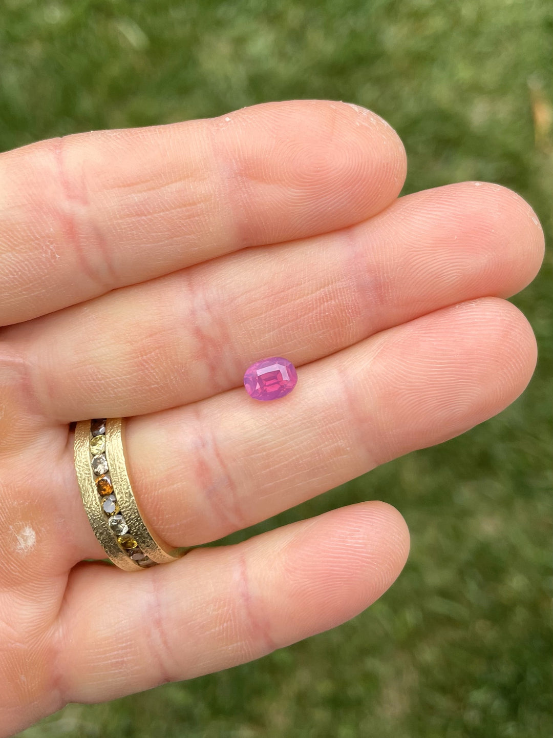 A 1.53-carat reddish purple and pink sapphire with opalescent glow displayed on fingertips, showcasing its vivid color and oval cut brilliance.