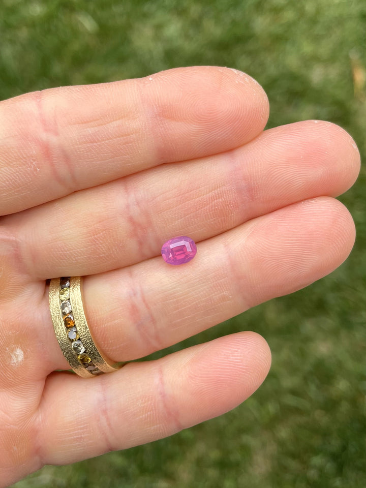 A 1.53-carat reddish purple and pink sapphire with opalescent glow displayed on fingertips, showcasing its vivid color and oval cut brilliance.