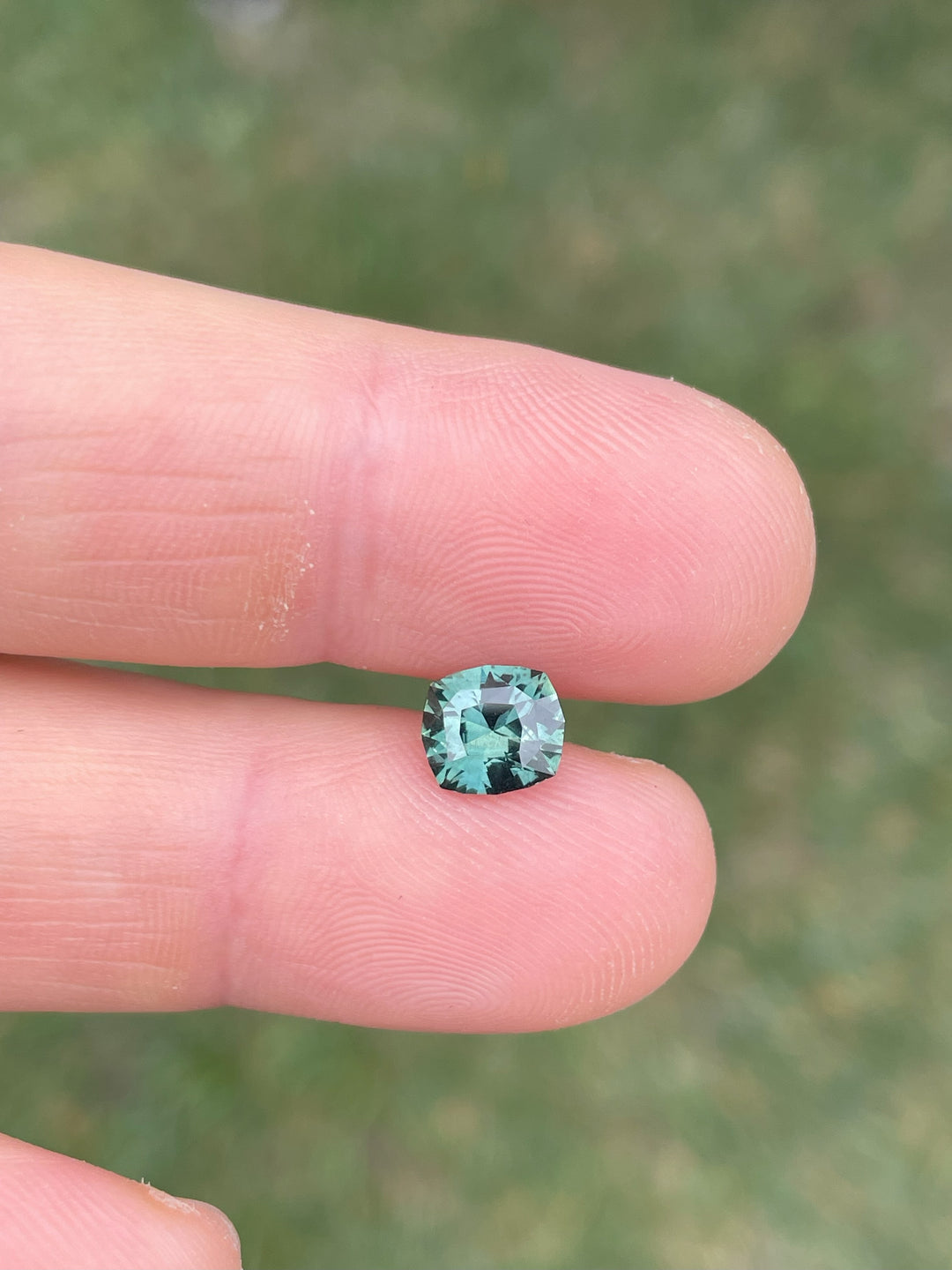 A close-up image of a 1.55-carat blue-green sapphire with a fancy mixed cut, displaying rich hues of blue and green. This unique gemstone is held between two fingers, showcasing its dimensions and intricate cut. Originating from Madagascar, this sapphire is untreated.