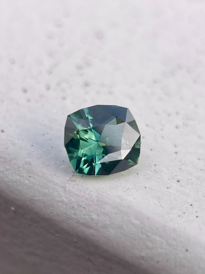 An angled view of a 1.55-carat blue-green sapphire from Madagascar. The fancy mixed cut reveals the gemstone's captivating color play and brilliance. The image highlights the stone's unique cut and untreated nature.