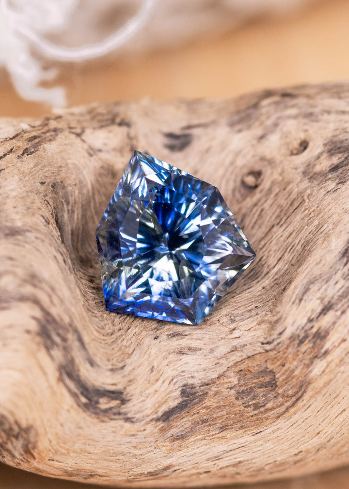 Loose blue and colorless parti sapphire from Sakaraha Madagascar resting on natural wood showing the custom geometric concave shield cut and sharp facet reflections.