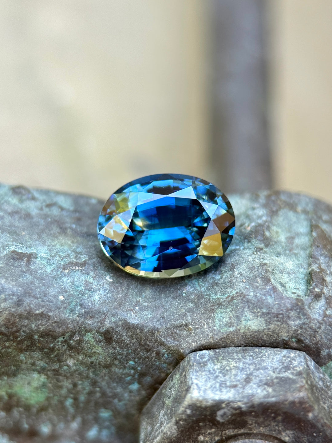 1.55 carat parti sapphire resting on textured stone surface, displaying vibrant yellow, teal, and blue color zoning in an oval mixed cut, untreated gemstone from Sakaraha, Madagascar.