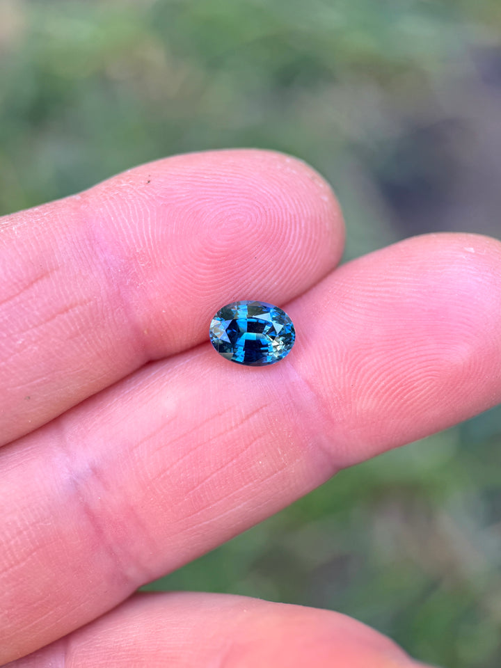 Close-up of 1.55 carat yellow teal blue parti sapphire resting on fingertips in daylight, highlighting its natural color transitions and faceted oval mixed cut of the untreated gem from Madagascar.