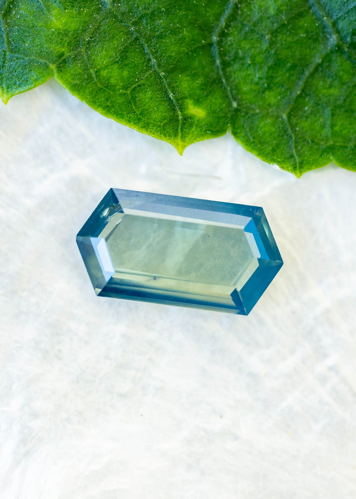 Natural 1.55 carat opalescent teal blue sapphire from Gombe, Nigeria, photographed beneath a green leaf on a pale surface. Its elongated hexagonal portrait cut displays a softly translucent seafoam center surrounded by saturated teal-blue step facets.