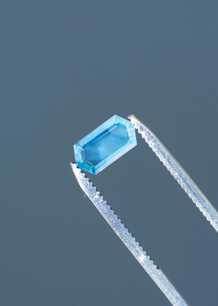 Close-up photograph of a 1.55ct untreated opalescent teal blue sapphire held in silver jeweler's tweezers. The elongated hexagonal portrait cut reveals crisp geometric edges, a broad central window and cool blue reflections.