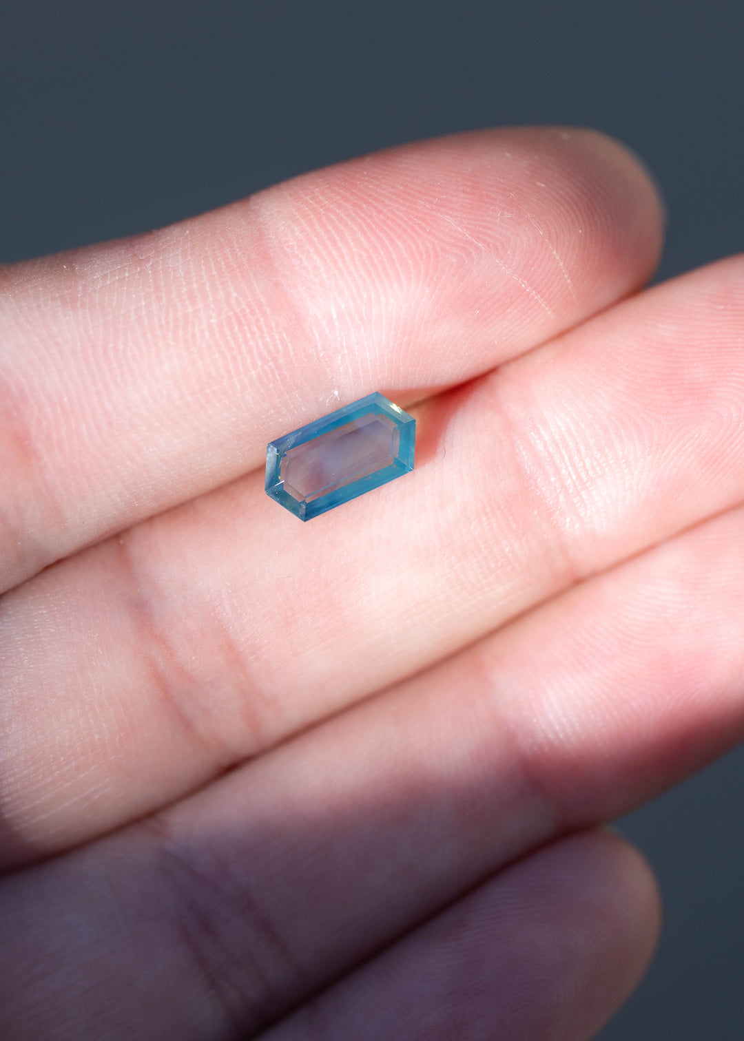 Untreated 1.55 carat opalescent teal blue sapphire from Gombe, Nigeria, photographed on fingers for scale. Its elongated hexagonal portrait cut measures 10.13 × 5.29 mm, with a shallow 2.31 mm depth.