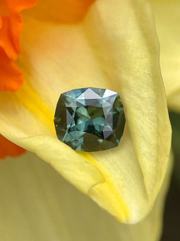 A 1.55-carat sapphire with a fancy mixed cut from Madagascar, featuring rich blue and green tones, displayed on a delicate yellow flower petal.