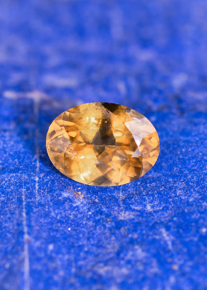 1.56 carat yellow orange Montana sapphire photographed on blue mineral background showing oval mix cut facets and fiery golden orange color from El Dorado Bar Deposit USA