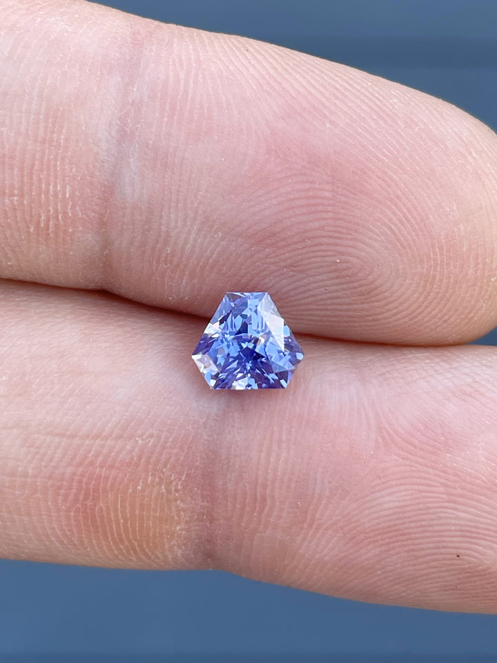 Close-up of a 1.56-carat lavender sapphire in a custom triangle brilliant cut, held between fingers. The gemstone showcases a mix of purple and pink hues with exceptional brilliance.