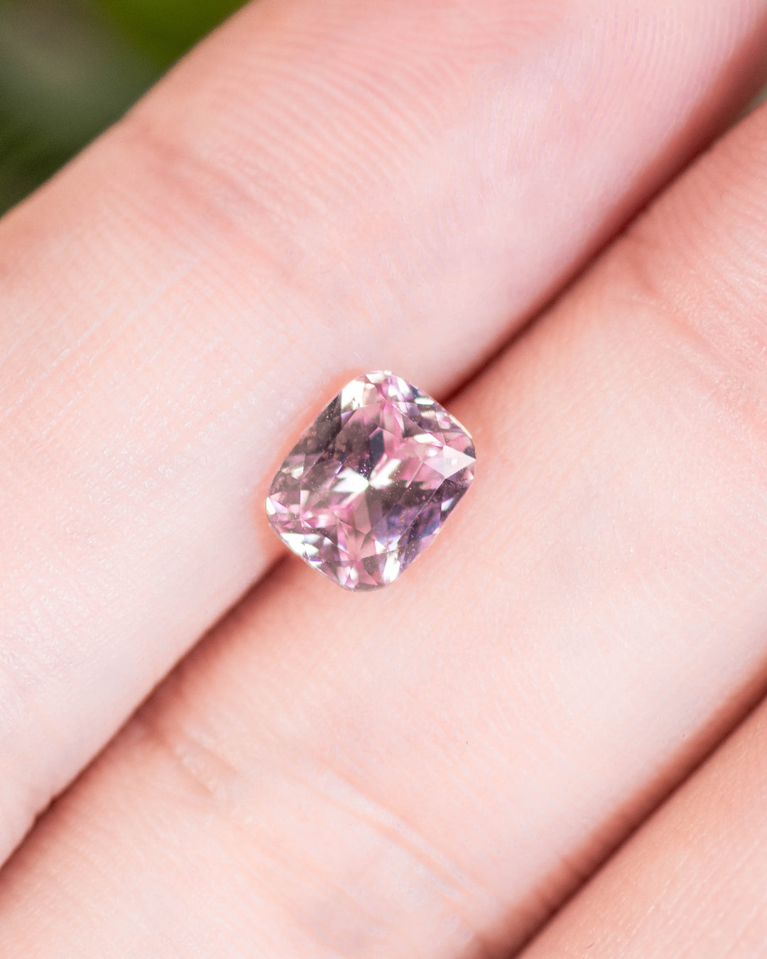1.57 carat pink and peach sapphire on fingertips under natural light, showing clarity and sparkle of the precision cushion cut, untreated gemstone from Sakaraha, Madagascar.