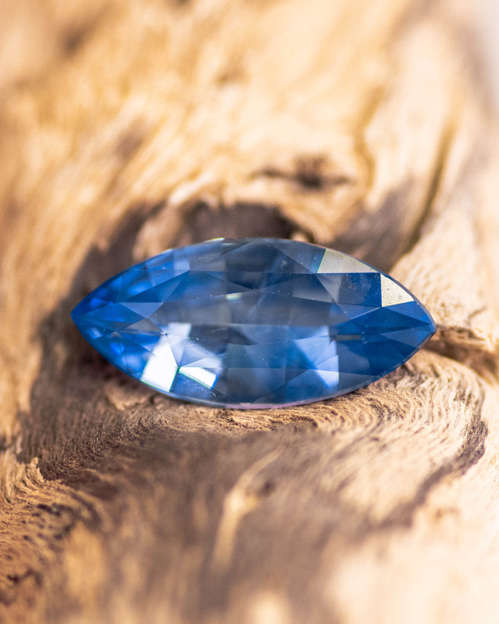 1.57CT vivid blue sapphire resting on textured wood surface, emphasizing rich blue saturation and fine faceting of the precision marquise mixed cut, unheated sapphire from Sakaraha, Madagascar.