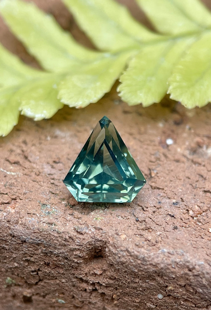 Close-up of a 1.57ct shield-cut sapphire featuring opalescent hues of yellow, teal, and green, highlighted by a step-cut design. The gemstone rests on a natural brown surface, with vibrant green foliage in the background.