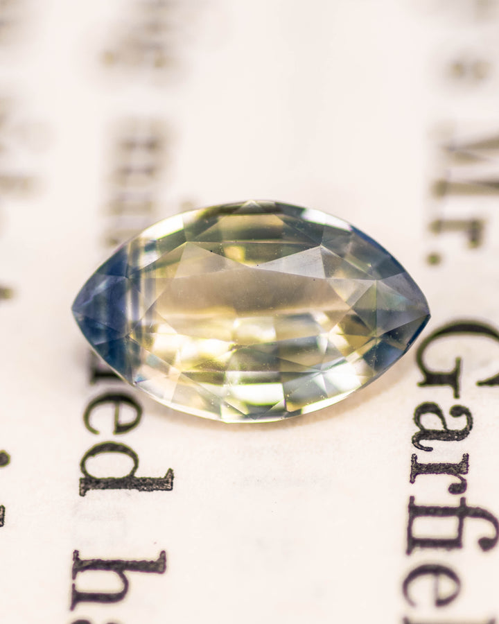 1.58 carat yellow and blue parti sapphire placed on text background, displaying intricate marquise mixed cut facets and glowing natural color transitions, unheated sapphire from Sakaraha, Madagascar.