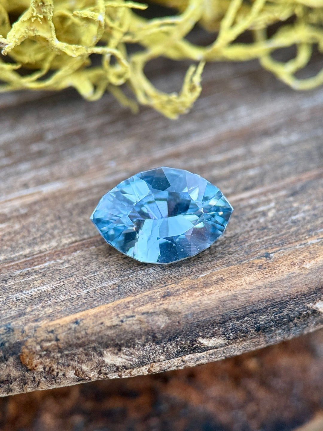 1.58 carat teal, blue, and green sapphire resting on rustic wood near yellow moss, showing natural brilliance and color zoning in a precision marquise mixed cut, untreated sapphire from Sakaraha, Madagascar.