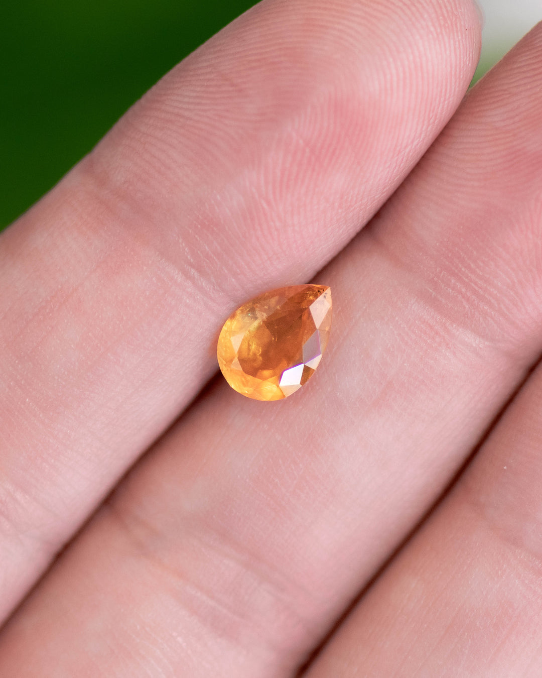 Close-up of 1.59 carat orange sapphire resting on fingertips, revealing warm yellow and red-orange color transitions and the sparkle of the modified pear brilliant cut, untreated sapphire from Umba River, Tanzania.