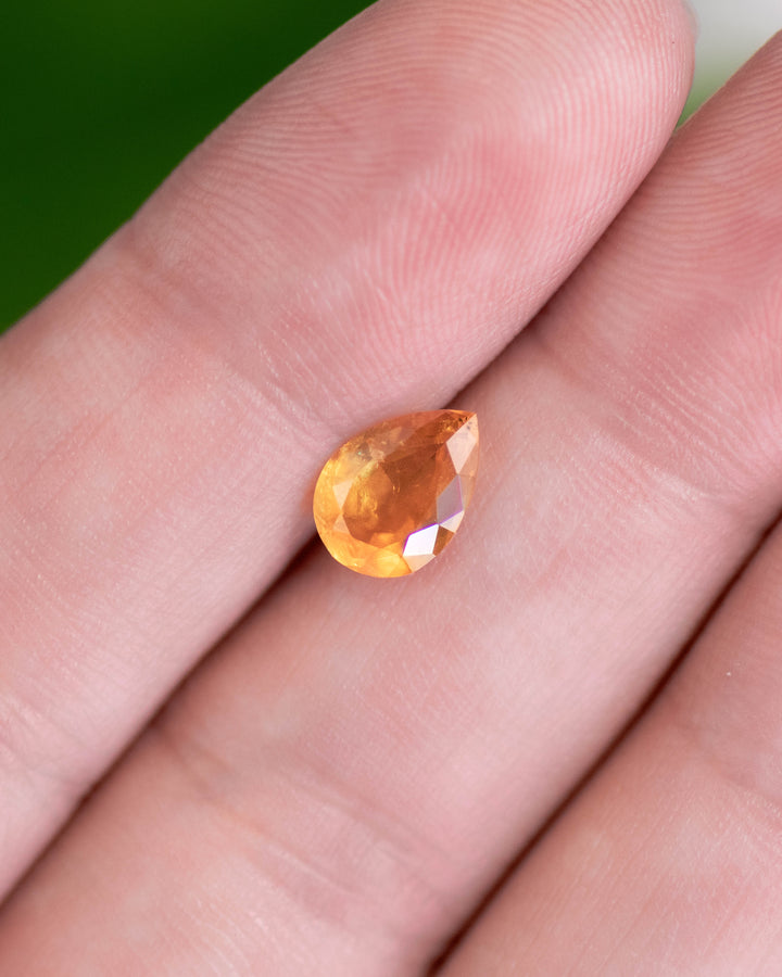 Close-up of 1.59 carat orange sapphire resting on fingertips, revealing warm yellow and red-orange color transitions and the sparkle of the modified pear brilliant cut, untreated sapphire from Umba River, Tanzania.