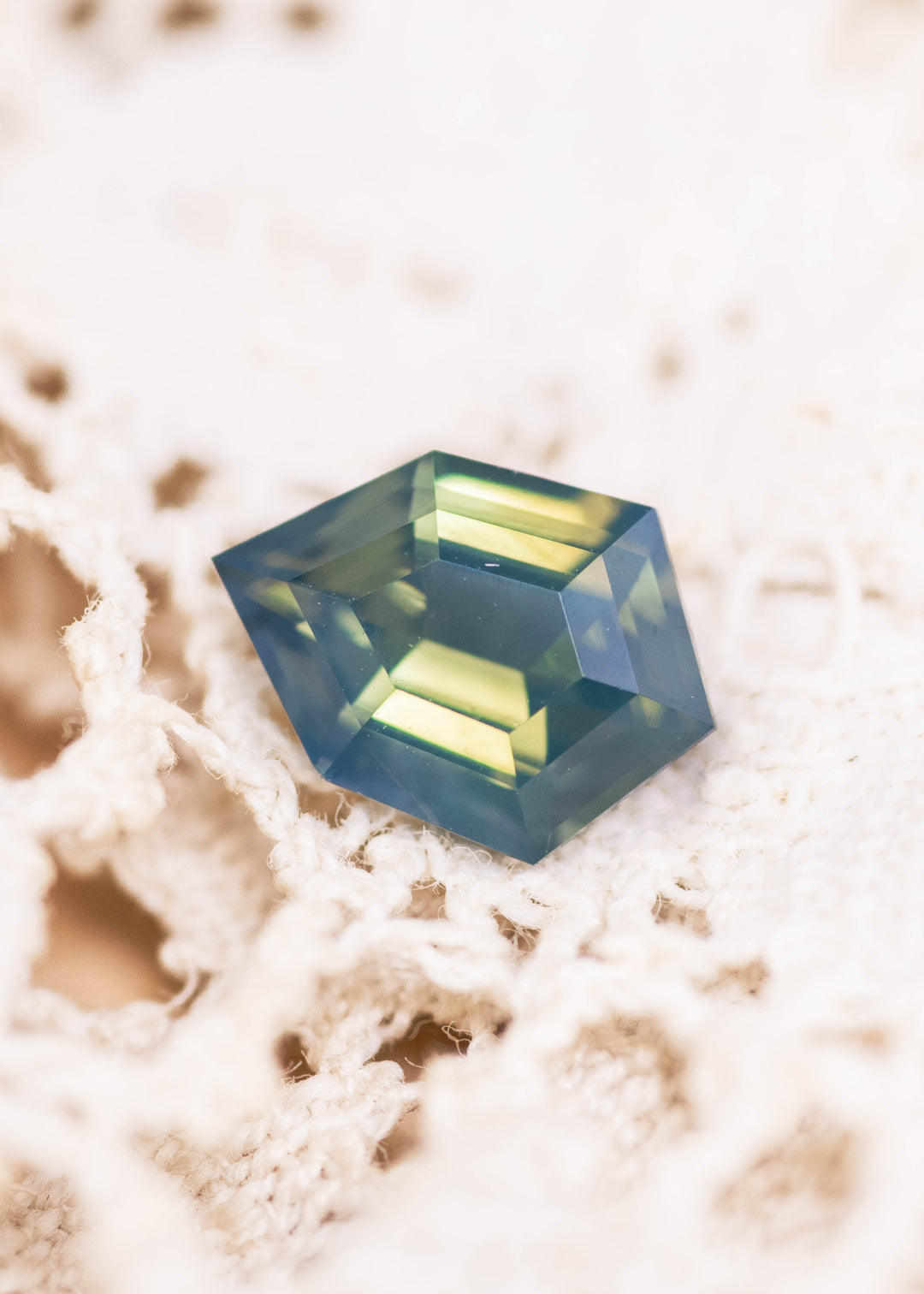Loose opalescent greenish teal blue sapphire from Diego, Madagascar shown face-up on textured lace, highlighting the shield step cut and broad step facets.